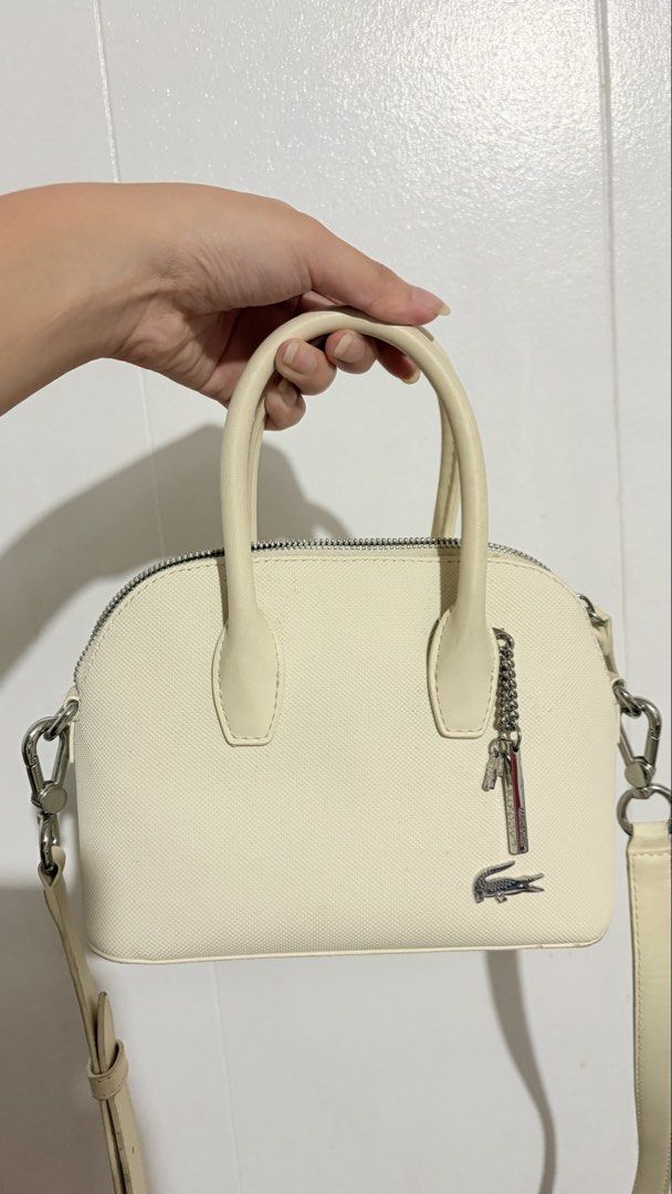 Lacoste Mini Daily Lifestyle Bugatti Strap Bag (Category: Cross-body Bags) located in Baguio. Submitted by meiji50 (Listing ID 1469930651, Image 1). Description: - Brand: Lacoste - Material: Cream textured fabric with black interior lining - Hardware: Silver-tone with zipper closure - Features: Twin top handles, detachable shoulder strap, front crocodile logo plate, branded metal charm, and an interior slip pocket - Condition: Minor wear on the e