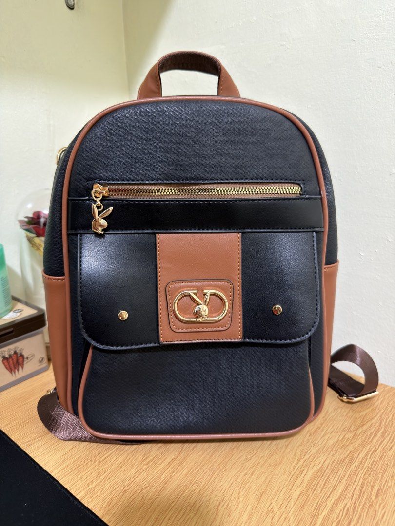 Playboy Bunny Two-Tone Black and Brown Leatherette Backpack (Category: Backpacks) located in Manila. Submitted by renee_12 (Listing ID 1469803769, Image 1). Description: - Color & Material: Black textured body with brown leatherette trim - Hardware: Gold-toned accents and interlocking metal detail on front flap - Charm: Zipper pull with a dangling gold Playboy bunny charm - Straps: Top carry handle and adjustable brown shoulder straps - Quality: 