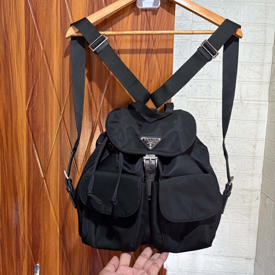 Prada Black Nylon Backpack (Category: Backpacks) located in Carmen. Submitted by ar_thrift00 (Listing ID 1469762693, Image 1). Description: - Brand: Prada - Material: Lightweight black nylon with silver-tone branded hardware - Design: Classic enamel triangle logo on the front flap - Closure: Drawstring main compartment with secure buckle strap - Storage: Dual exterior front pockets with flaps for easy access - Straps: Adjustable black webbed shou