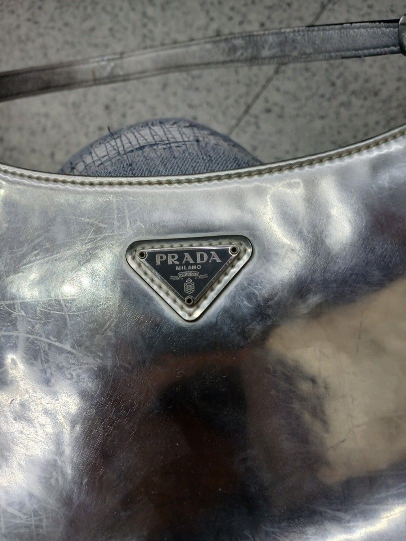 Prada Silver Shoulder Bag (Category: Luxury Bags & Wallets) located in Manila. Submitted by zackthrift16 (Listing ID 1469847834, Image 2). Description: - Brand: Prada - Color: Metallic silver - Style: Curved shoulder bag with single strap - Features: Front enamel triangle logo plaque, magnetic snap closure, and interior zip pocket with Prada Made in Italy metal plaque - Condition: Noticeable scuffs and surface wear on the metallic finish; hardwar