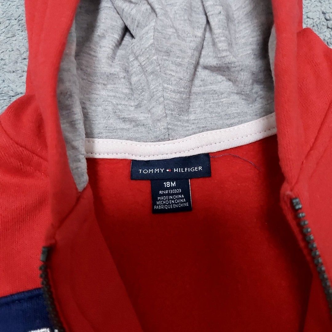 Tommy Hilfiger 18M Red Hooded Zip Jacket (Category: Babies & Kids Fashion) located in Caloocan City. Submitted by epicshark.da6ff7 (Listing ID 1469892792, Image 2). Description: - Brand: Tommy Hilfiger - Size: 18 Months - Color: Vibrant red with navy blue and white accents - Design: Contrast navy chest band with white embroidered lettering - Features: Full front zip closure, grey hood lining, and two front pockets - Condition: Clean fabric and br