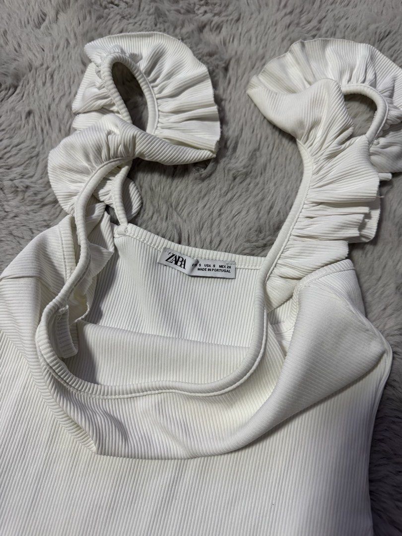 Zara Ruffled Strap Ribbed Bodysuit White (Category: Sleeveless) located in Calamba. Submitted by cheriiv (Listing ID 1469883391, Image 3). Description: - Brand: Zara - Style: White ribbed bodysuit with square neckline and thick ruffle straps - Size: Small - Details: Snap button closure at the bottom, made in Portugal - Material: Stretchy cotton blend.