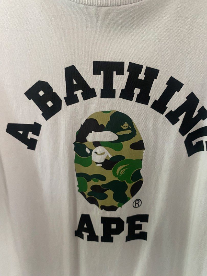 A Bathing Ape Camo Logo White T-Shirt (Category: Tshirts & Polo Shirts ) located in Manila. Submitted by upcycleutopia_ (Listing ID 1469982134, Image 2). Description: - Brand: A Bathing Ape (BAPE) - Color: Clean Solid White - Size: Large - Design: Large arched text logo with classic green camo ape head on the chest - Details: Short sleeves, crew neck, tag attached at inner collar - Material: 100% Cotton - Condition: good -issue scratch on sleeve 