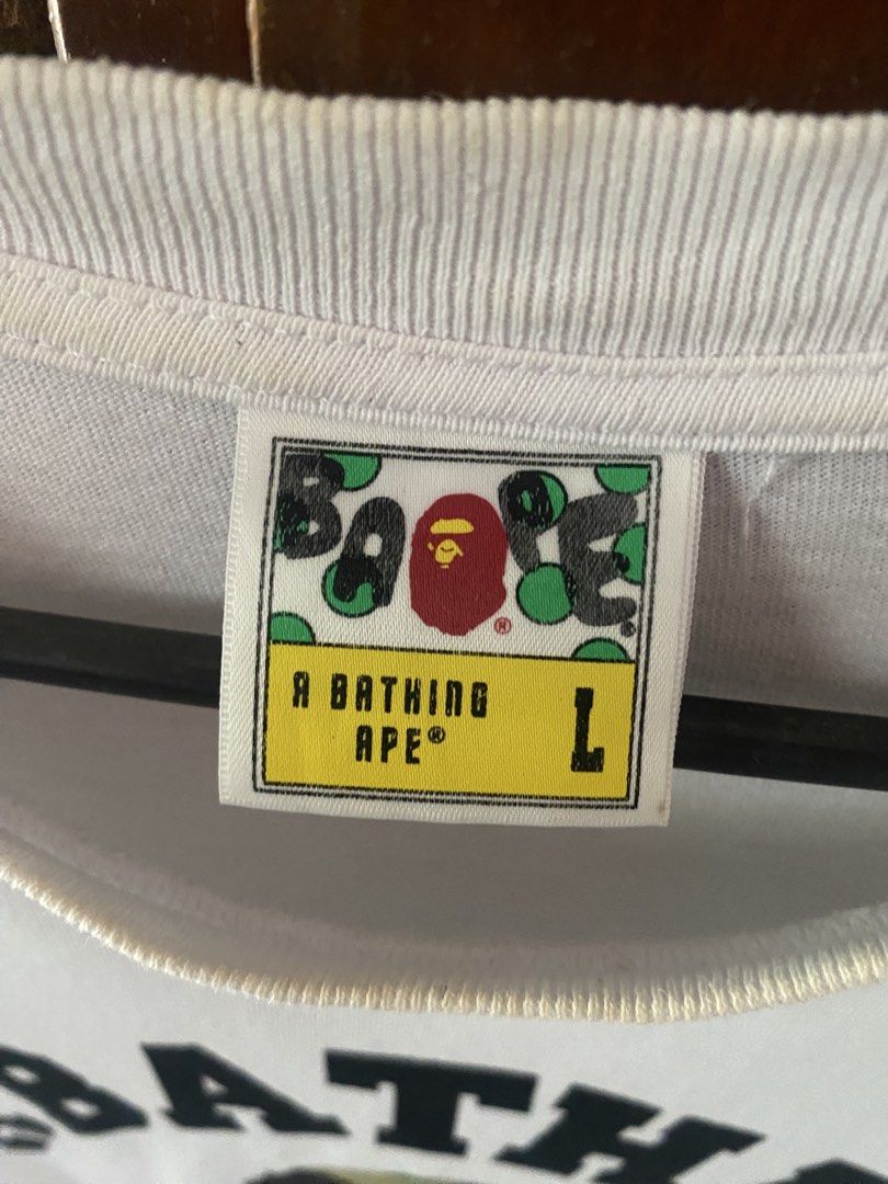 A Bathing Ape Camo Logo White T-Shirt (Category: Tshirts & Polo Shirts ) located in Manila. Submitted by upcycleutopia_ (Listing ID 1469982134, Image 3). Description: - Brand: A Bathing Ape (BAPE) - Color: Clean Solid White - Size: Large - Design: Large arched text logo with classic green camo ape head on the chest - Details: Short sleeves, crew neck, tag attached at inner collar - Material: 100% Cotton - Condition: good -issue scratch on sleeve 
