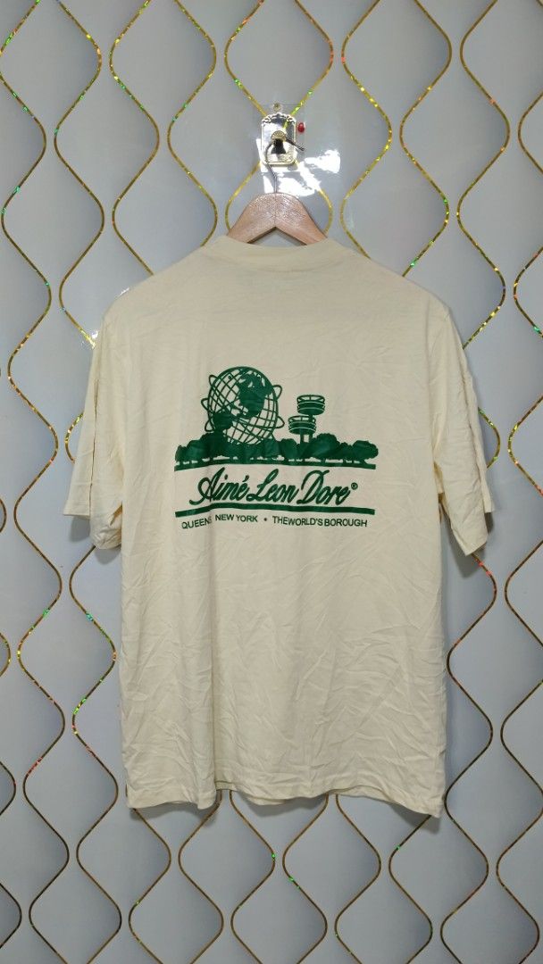Aimé Leon Dore Queens World's Borough Tee (Category: Tshirts & Polo Shirts ) located in Manila. Submitted by zariyah.1986 (Listing ID 1469950106, Image 3). Description: - Brand: Aimé Leon Dore - Color: Cream with green graphic print - Design: Small chest logo and large back graphic featuring the Queens Unisphere and landmarks - Details: Crew neck, short sleeves, clean collar - Measurements: 21.5 inches pit to pit, 28.5 inches length - Condition: 