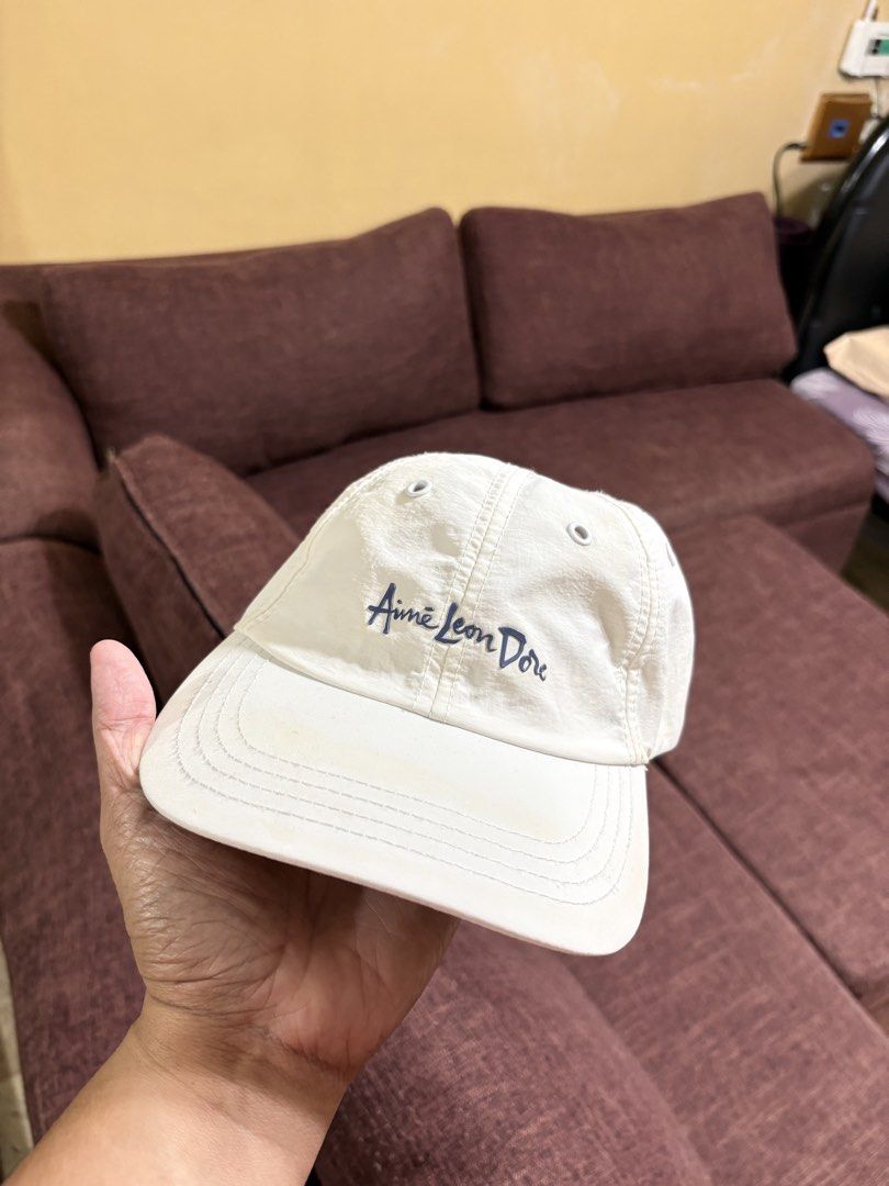Aimé Leon Dore White Cap (Category: Caps & Hats) located in Manila. Submitted by breezy09breezy (Listing ID 1469987920, Image 2). Description: - Brand: Aimé Leon Dore - Color: White with blue cursive embroidered logo - Closure: Adjustable black strap with branded tag - Design: Classic six-panel with ventilation eyelets - Condition: Good, with minor yellowish discoloration on the interior sweatband from wear.