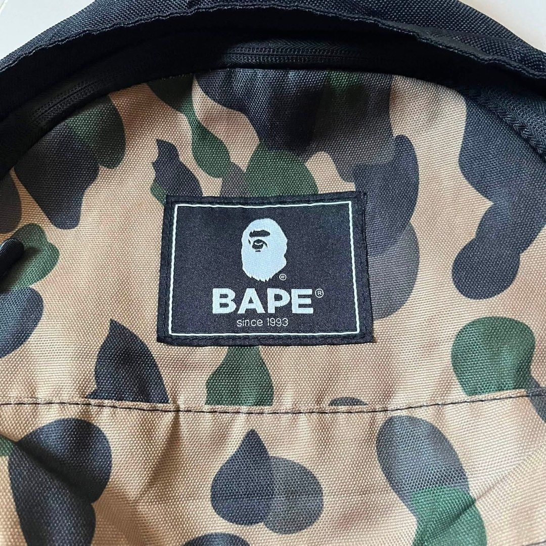 BAPE Camo Backpack with White Side Branding (Category: Backpacks) located in Toledo. Submitted by collective_stuff (Listing ID 1469956840, Image 2). Description: e-MOOK A BATHING APE 2021 SUMMER COLLECTION - Classic black backpack featuring iconic BAPE camo panel - Bold white BAPE text printed along both side panels - Durable canvas material with sturdy construction for daily use - Comfortable black shoulder straps with adjustable buckles - Ideal