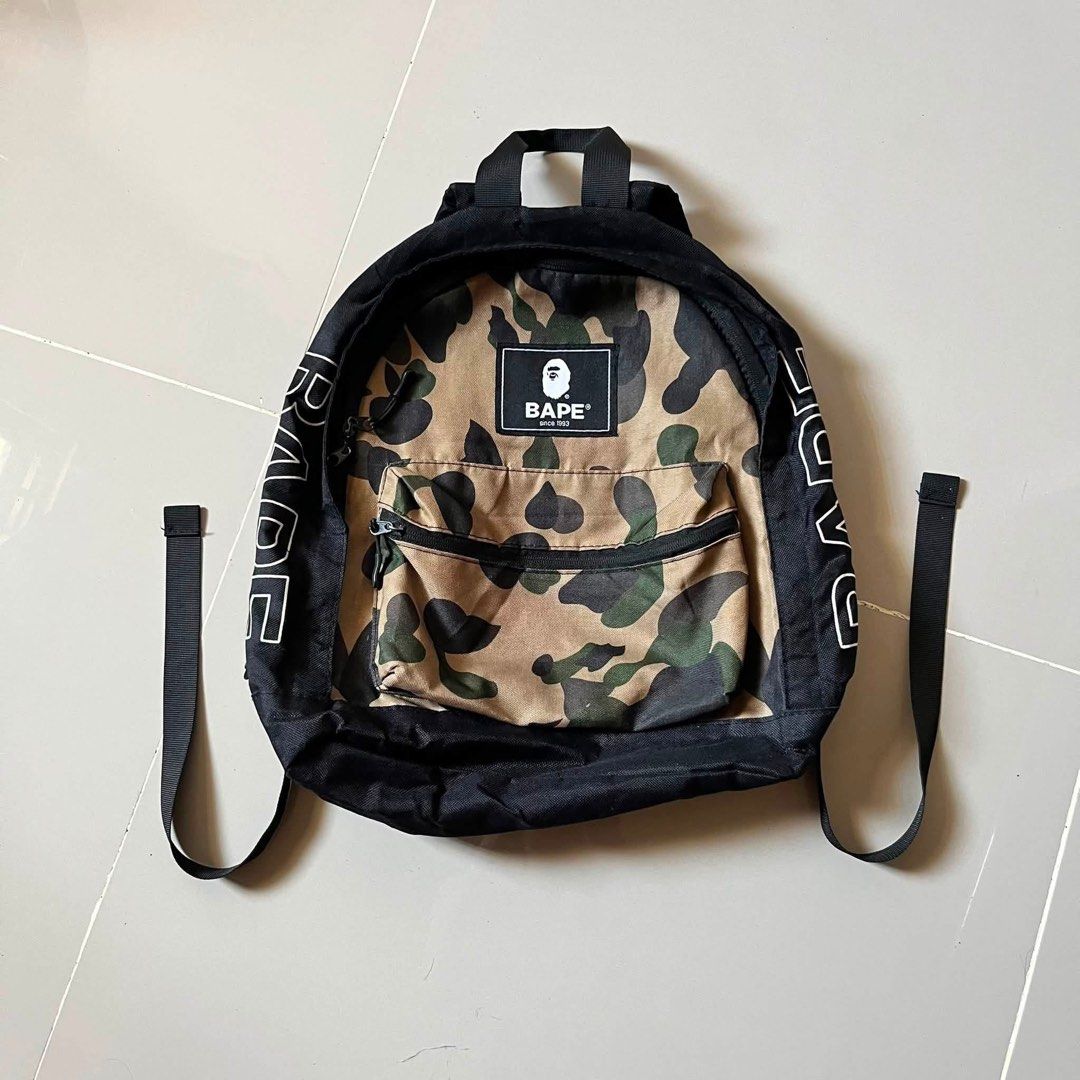 BAPE Camo Backpack with White Side Branding (Category: Backpacks) located in Toledo. Submitted by collective_stuff (Listing ID 1469956840, Image 1). Description: e-MOOK A BATHING APE 2021 SUMMER COLLECTION - Classic black backpack featuring iconic BAPE camo panel - Bold white BAPE text printed along both side panels - Durable canvas material with sturdy construction for daily use - Comfortable black shoulder straps with adjustable buckles - Ideal