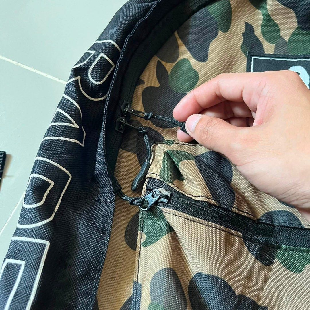 BAPE Camo Backpack with White Side Branding (Category: Backpacks) located in Toledo. Submitted by collective_stuff (Listing ID 1469956840, Image 3). Description: e-MOOK A BATHING APE 2021 SUMMER COLLECTION - Classic black backpack featuring iconic BAPE camo panel - Bold white BAPE text printed along both side panels - Durable canvas material with sturdy construction for daily use - Comfortable black shoulder straps with adjustable buckles - Ideal
