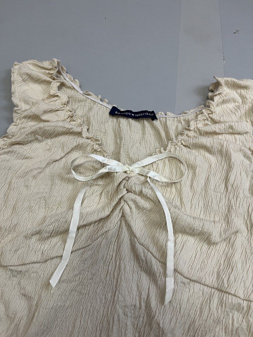 Brandy Melville Cream Ruffled Bow Tank Top (Category: Sleeveless) located in Mabalacat. Submitted by aas_thriftfings (Listing ID 1470105657, Image 2). Description: - Brand: Brandy Melville - Color: Cream - Fabric: Textured crinkle material (55% cotton, 42% polyester, 3% spandex) - Details: Lettuce trim edges, scoop neck, and a cute little white ribbon at the center front bust - Condition: Excellent, no flaws spotted - Care: Machine wash max 30 de