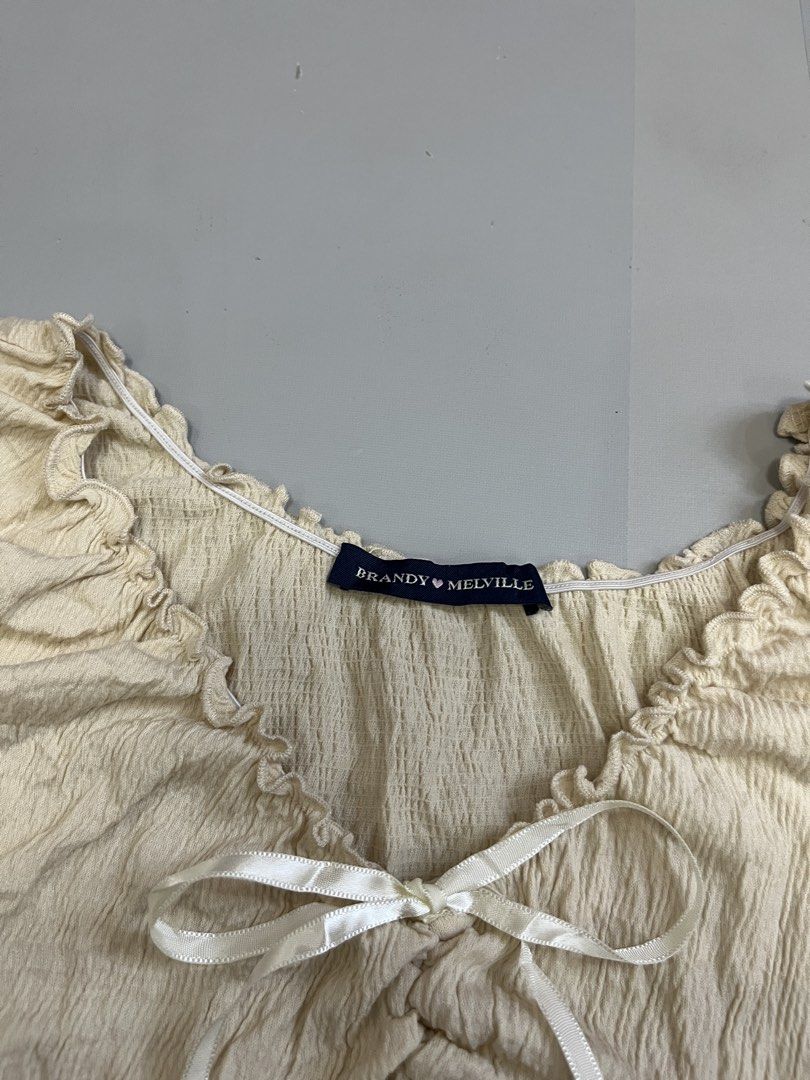 Brandy Melville Cream Ruffled Bow Tank Top (Category: Sleeveless) located in Mabalacat. Submitted by aas_thriftfings (Listing ID 1470105657, Image 3). Description: - Brand: Brandy Melville - Color: Cream - Fabric: Textured crinkle material (55% cotton, 42% polyester, 3% spandex) - Details: Lettuce trim edges, scoop neck, and a cute little white ribbon at the center front bust - Condition: Excellent, no flaws spotted - Care: Machine wash max 30 de