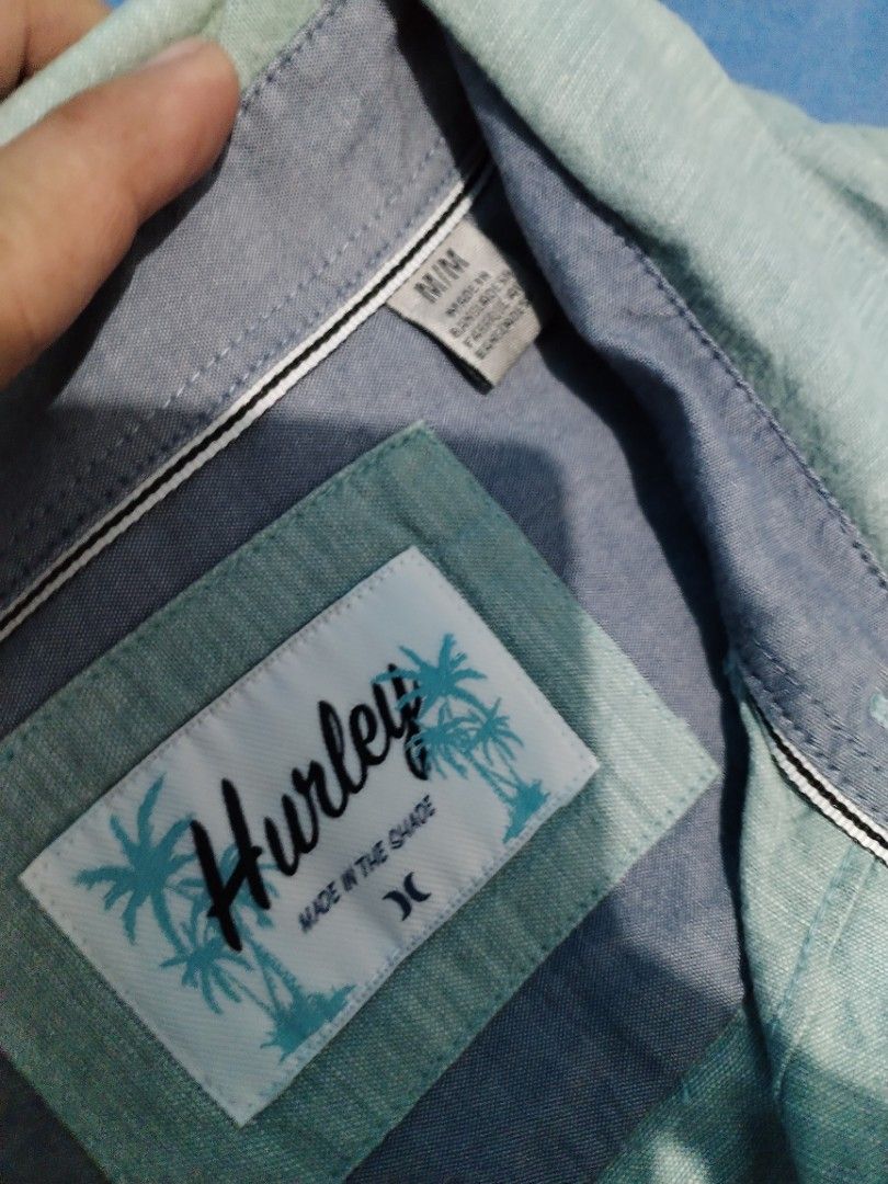 Hurley Men's Short Sleeve Button-Down Shirt Size M (Category: Tshirts & Polo Shirts ) located in Manila. Submitted by solebrothers2826 (Listing ID 1470106403, Image 3). Description: - Brand: Hurley - Style: Short sleeve button-down shirt - Size: Medium - Color: Light blue with subtle textured pattern and contrast inner collar trim - Details: Left chest pocket with black logo tab, palm tree graphic patch near the bottom hem - Material: Lightweight
