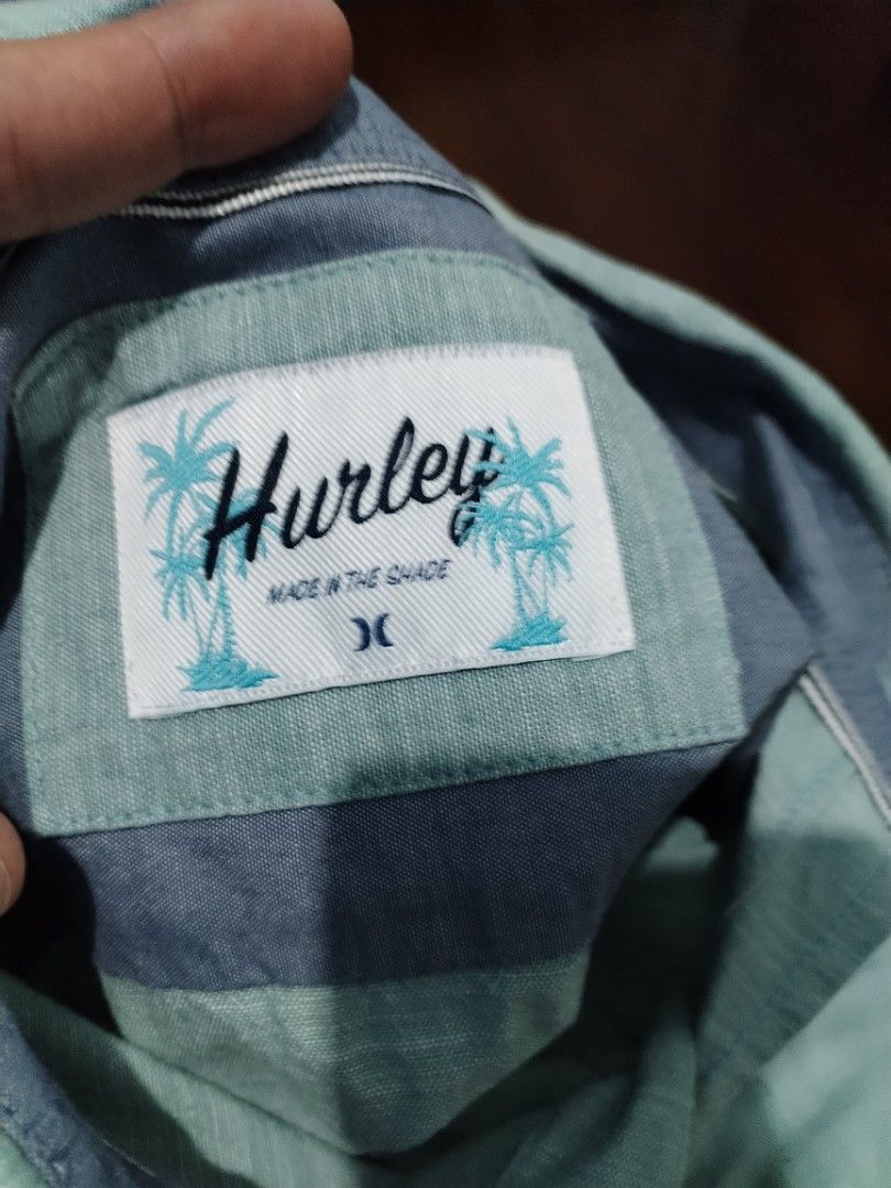 Hurley Men's Short Sleeve Button-Down Shirt Size M (Category: Tshirts & Polo Shirts ) located in Manila. Submitted by solebrothers2826 (Listing ID 1470106403, Image 2). Description: - Brand: Hurley - Style: Short sleeve button-down shirt - Size: Medium - Color: Light blue with subtle textured pattern and contrast inner collar trim - Details: Left chest pocket with black logo tab, palm tree graphic patch near the bottom hem - Material: Lightweight