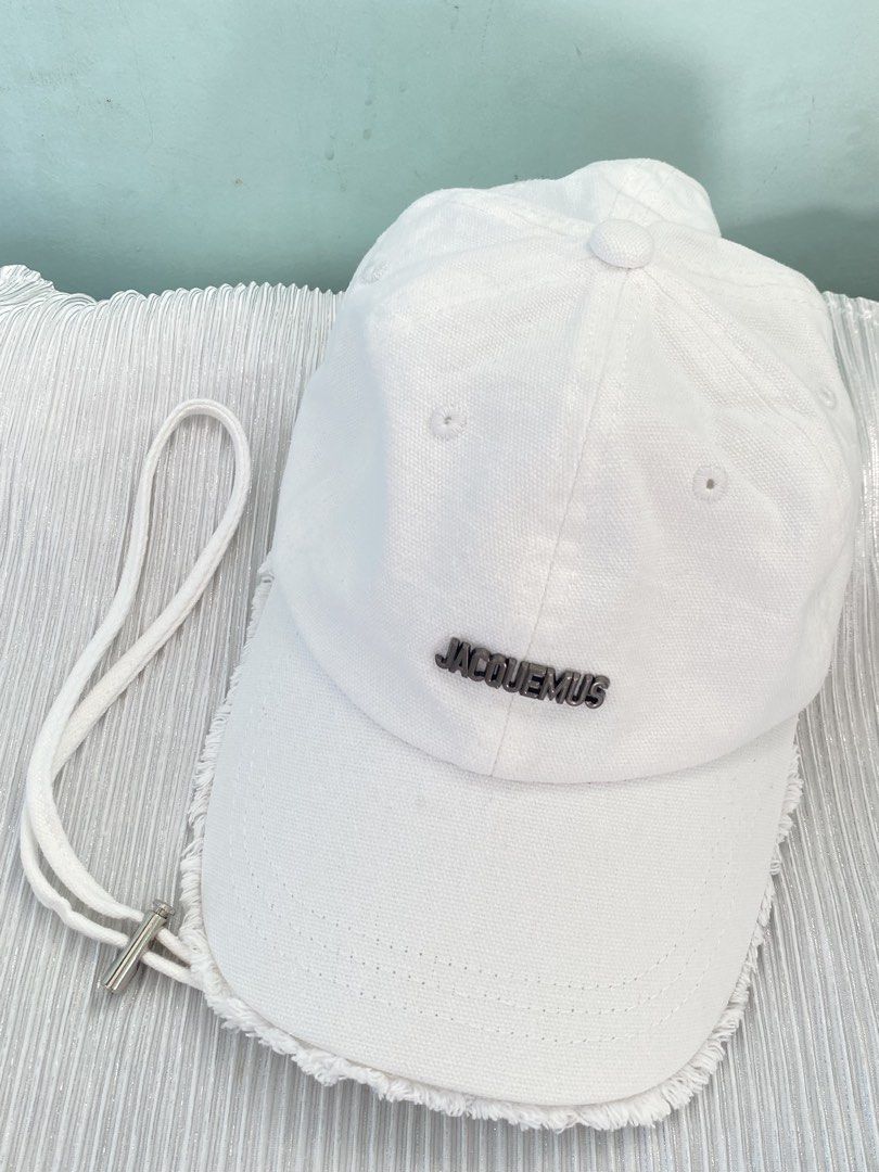 Jacquemus White Cotton Baseball Cap (Category: Caps & Hats) located in Tabaco. Submitted by camila_p (Listing ID 1470048626, Image 2). Description: - Authentic Jacquemus baseball cap in crisp white cotton canvas - Features a sleek metal JACQUEMUS lettering logo on the front - Trendy raw frayed edge detailing on the brim - Adjustable back strap with a branded silver-tone metal buckle - Interior includes official black and white brand tags (Made in
