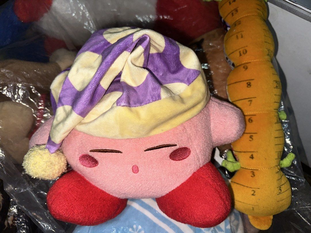 Kirby with Sleeping Cap with papertag (Category: Plushies & Soft Toys) located in Pasig City. Submitted by katplush_superlove (Listing ID 1470043021, Image 3). Description: - Official-style cute Kirby plush figure in a sleeping pose - Features a cozy purple and yellow patterned sleeping cap with a pom-pom - Made with ultra-soft, huggable pink fabric with embroidered closed eyes and red feet - Tag attached near the side, great for collectors - Per