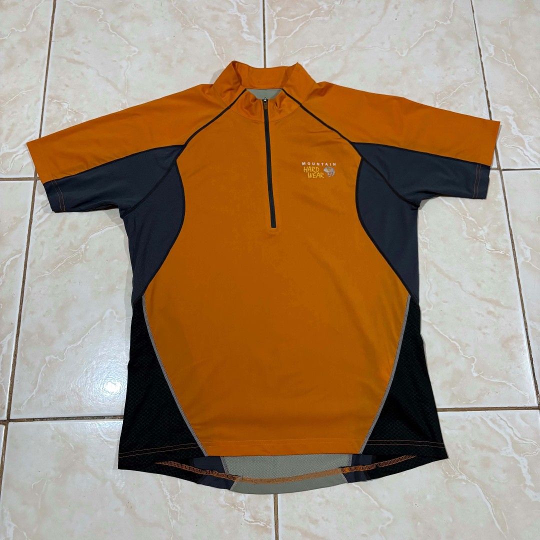 Mountain Hardwear Orange (Category: Activewear) located in El Salvador. Submitted by pretiezt2001 (Listing ID 1470047317, Image 1). Description: - Brand: Mountain Hardwear - Color: Bright orange and dark grey - Design: Short sleeves, half-zip front, and high collar - Breathability: Mesh fabric panels on shoulders and back yoke - Safety: Reflective piping details along seams - Branding: Clear logo printed on the chest - Ideal for: Outdoor rides, r