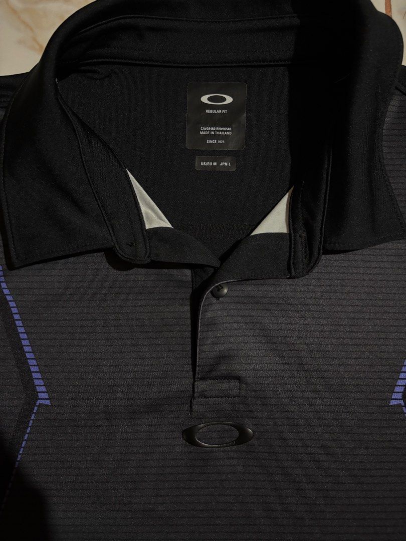 Oakley Black Regular Fit Polo Shirt (Category: Tshirts & Polo Shirts ) located in Dumaguete. Submitted by revillahar03975 (Listing ID 1469982442, Image 2). Description: - Brand: Oakley - Style: Regular fit polo shirt with short sleeves and standard collar - Color: Black with purple geometric stripe accents - Details: Single button placket with signature Oakley ellipse hardware loop - Size: L .