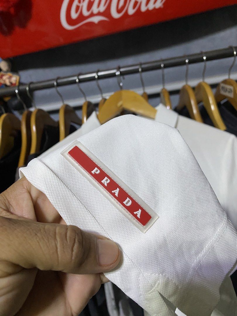 Prada White Polo Shirt (Category: Luxury Apparel) located in Santa Ana. Submitted by tunes_vtg (Listing ID 1470013220, Image 3). Description: - Prada polo shirt in clean white cotton pique - Classic collar with branded mother-of-pearl buttons - Signature enameled metal triangle logo on the chest - Red Prada tag on the lower hem and woven interior collar label - Short sleeves with a standard, comfortable fit - Timeless staple piece, perfect for sm