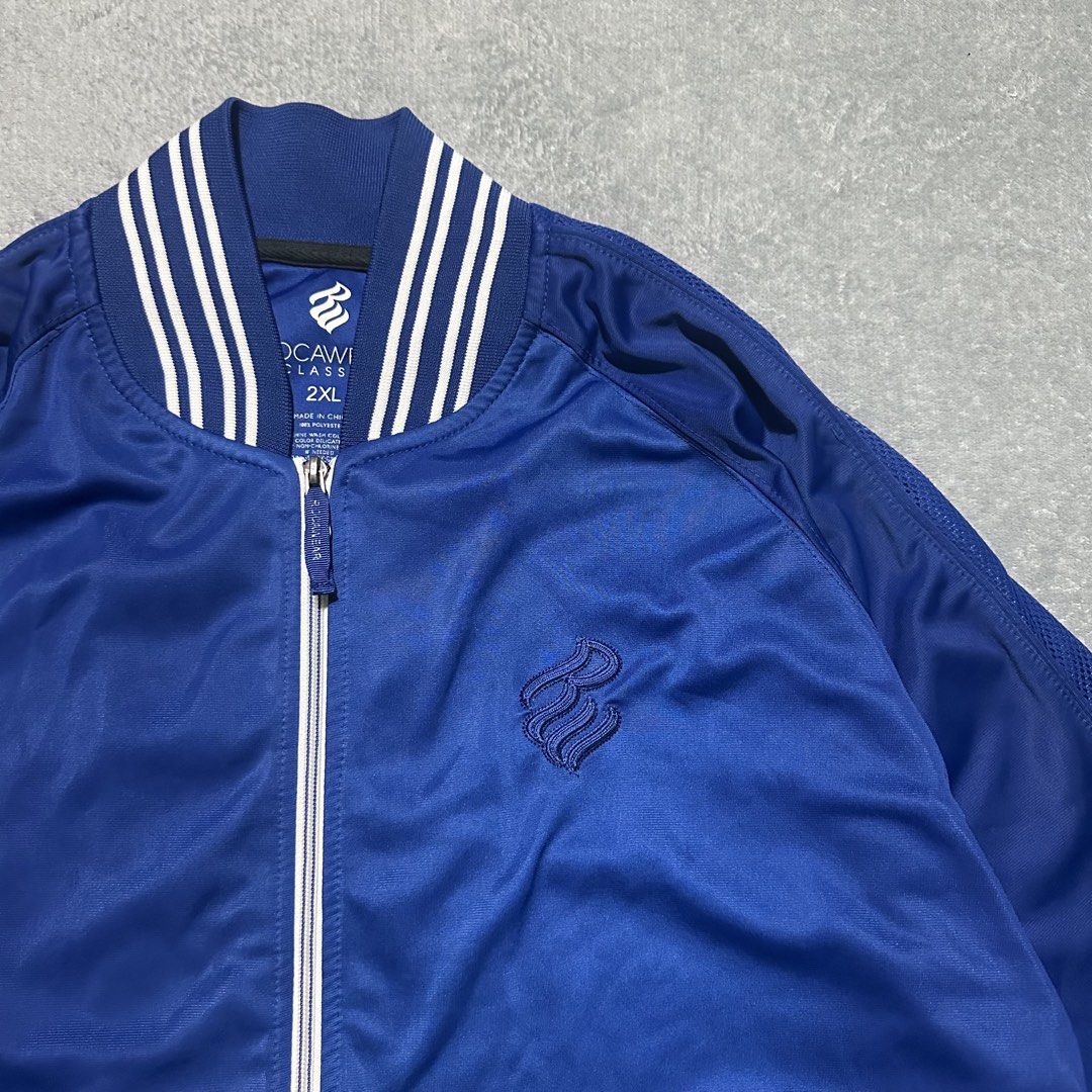 Rocawear Classic Blue Track Jacket 2XL (Category: Coats, Jackets and Outerwear) located in Villaverde. Submitted by bakatzy_secondhand_garments (Listing ID 1470093744, Image 3). Description: - Brand/Model: Rocawear Classic Track Jacket - Color: Royal Blue - Size: 2XL - Design: Full zip front, striped ribbed collar, cuffs, and hem - Features: Two front pockets with white piping, subtle embroidered chest logo - Style: Classic athletic streetwear, p