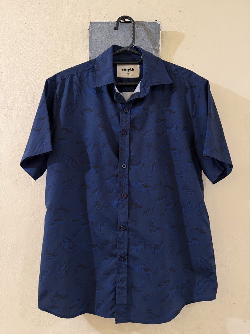 Smyth Blue Leaf Print Short Sleeve Shirt (Category: Tshirts & Polo Shirts ) located in Las Pinas. Submitted by rareriroruru (Listing ID 1470056119, Image 1). Description: - Brand/Style: Smyth navy blue short sleeve button-up shirt - Pattern: Dark leaf or feather print - Size: XS - Length: 26 in - Width: 20 in - Material: Lightweight fabric, ideal for warm weather - Condition: Good casual piece with minor fabric wear spots near the lower back hem 