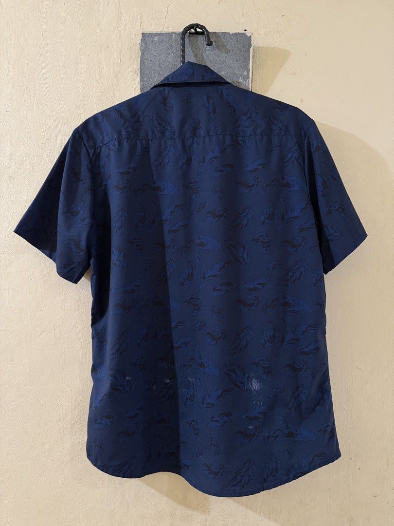Smyth Blue Leaf Print Short Sleeve Shirt (Category: Tshirts & Polo Shirts ) located in Las Pinas. Submitted by rareriroruru (Listing ID 1470056119, Image 2). Description: - Brand/Style: Smyth navy blue short sleeve button-up shirt - Pattern: Dark leaf or feather print - Size: XS - Length: 26 in - Width: 20 in - Material: Lightweight fabric, ideal for warm weather - Condition: Good casual piece with minor fabric wear spots near the lower back hem 