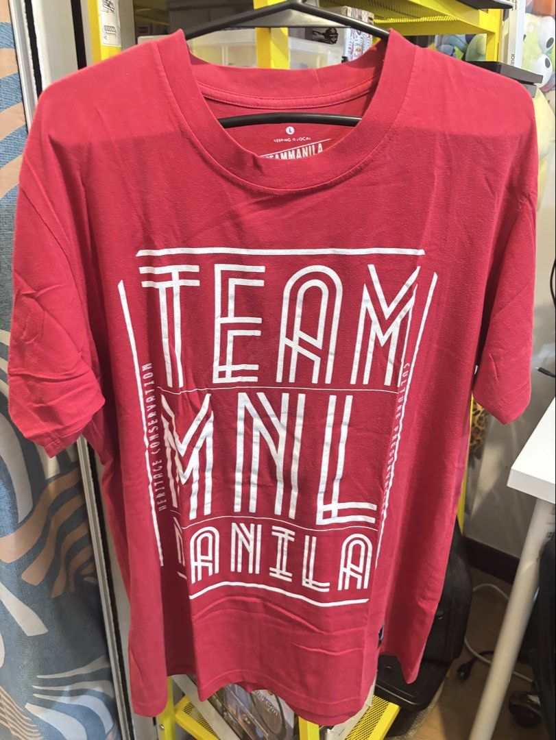 Team Manila Red Graphic T-Shirt (Category: Tshirts & Polo Shirts ) located in Navotas City. Submitted by psgvv (Listing ID 1470090849, Image 2). Description: - Brand: Team Manila - Color: Vibrant red - Size: L (tagged on inner collar) - Design: White block text print with border lines and side details on chest - Details: Crew neckline, short sleeves, and woven fabric label near bottom hem - Back: Clean, solid red.