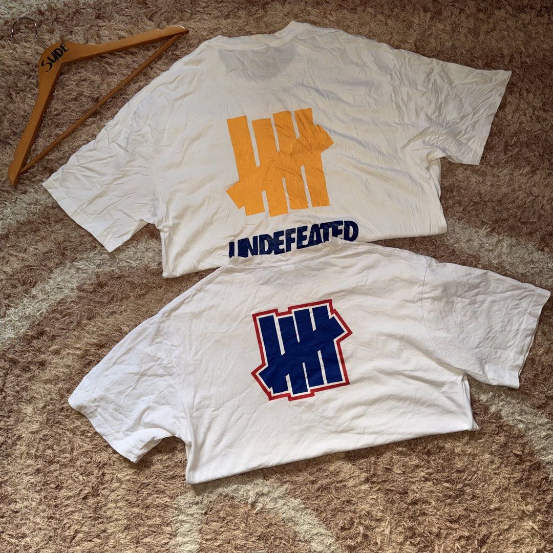 Undefeated White Graphic T-Shirt Bundle (Category: Tshirts & Polo Shirts ) located in Daraga. Submitted by sudeboy031092 (Listing ID 1470057380, Image 2). Description: - Bundle of 2 white Undefeated graphic tees - Material: 100% comfortable cotton - Tee 1: Small blue front logo with a large yellow and blue back logo - Tee 2: Red-bordered blue chest logo with a matching large back logo.