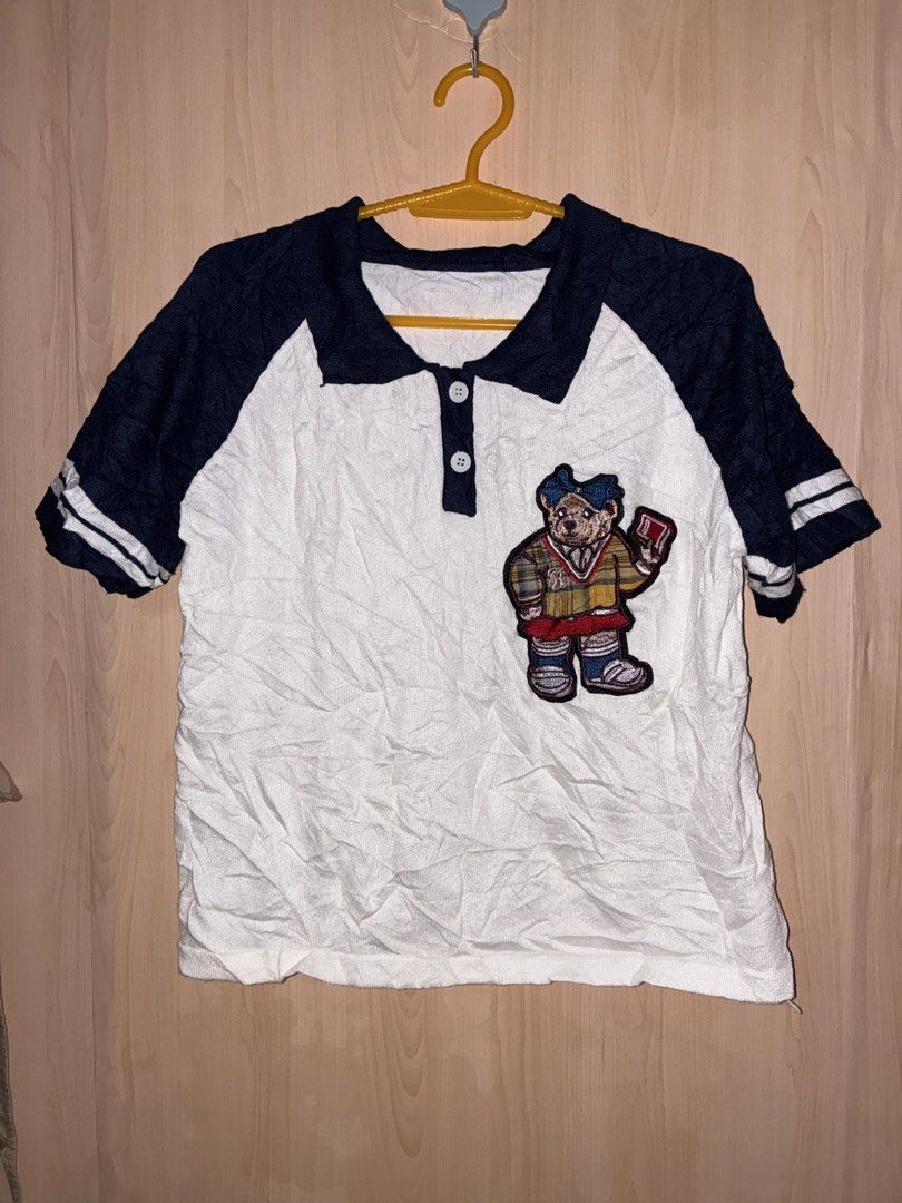White and Navy Polo Shirt with Bear Patch (Category: Blouses) located in Placer. Submitted by talaalond61563 (Listing ID 1470137386, Image 1). Description: - White short sleeve polo shirt with dark navy raglan sleeves - Features a large embroidered teddy bear patch on the chest - Classic collar with a 2-white-button placket - Stylish white stripe details on the sleeve cuffs - Made from soft, comfortable knit fabric - Perfect for casual wear and e
