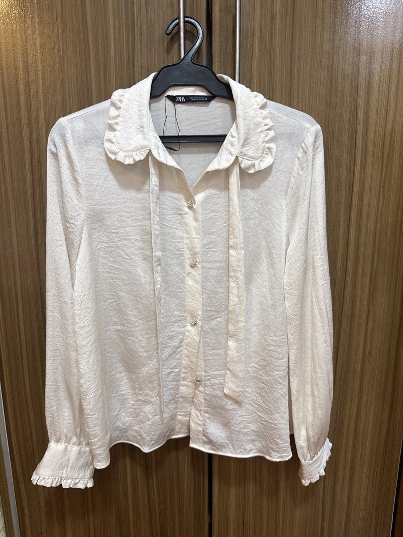 Zara White Ruffle Collar Blouse (Category: Blouses) located in Quezon City. Submitted by dropdeadprices (Listing ID 1470013046, Image 3). Description: - Brand: Zara - Condition: Brand new and never worn - Color: White - Details: Ruffle collar with neck tie string, button-up front with pearl buttons, ruffled cuffs - Fabric: Lightweight textured material.