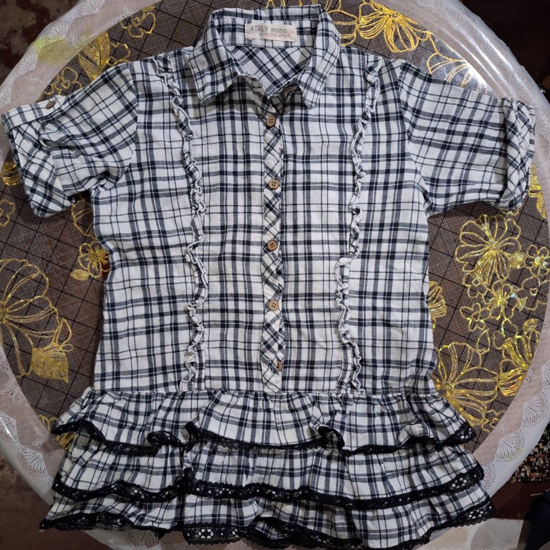 AIRLY momo plaid ruffle bluse/gingham tier (Category: Blouses) located in Pasig City. Submitted by zelzelly (Listing ID 1470289990, Image 1). Description: RFS: Decluterring, fits on me but too small for my arms. • xxs - xs size / 130 on tag 7 to 8yrs old kid size, but it fits on me when I wore this (xs) • AIRLY momo on tag (Japanese brand) Details: • Classic monochrome grid plaid (gingham-style). • Vertical ruffle panels framing the central butto