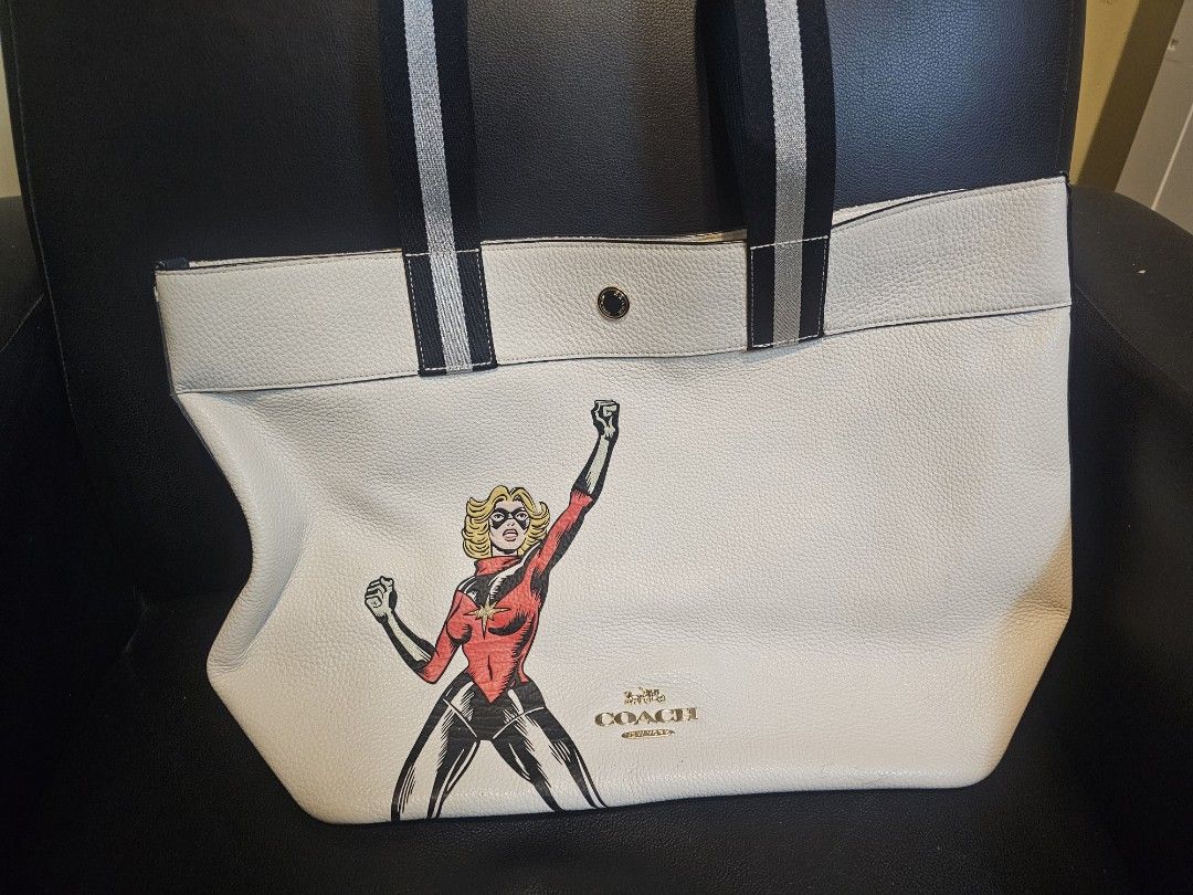 Coach Marvel Captain Marvel Tote Bag White (Category: Tote Bags) located in Las Pinas. Submitted by surplusxchange (Listing ID 1470168787, Image 1). Description: - Authentic Coach leather tote bag - Features a bold Captain Marvel graphic print on the front - Made from textured white pebble leather with black shoulder straps and red underside detail - Secure top snap closure - Brown fabric-lined interior with a zip pocket and Coach creed patch - I