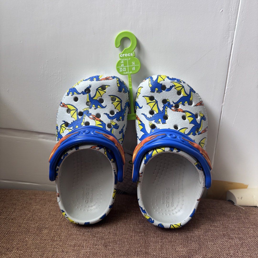 Crocs Classic Dragon Graphic Clog Toddler Size C6 (Category: Babies & Kids Fashion) located in Quezon City. Submitted by eshymarie (Listing ID 1470168491, Image 2). Description: - Item: Crocs Classic Dragon Graphic Clog for Toddlers - Size: C6 (EUR 22-23) for 13cm foot length - Design: White base with vibrant blue and yellow dragon print - Features: Blue pivoting heel strap with flame details on the sides - Condition: Brand new with original tags