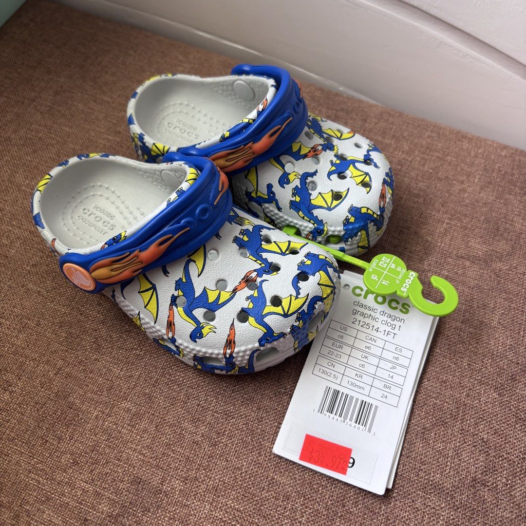 Crocs Classic Dragon Graphic Clog Toddler Size C6 (Category: Babies & Kids Fashion) located in Quezon City. Submitted by eshymarie (Listing ID 1470168491, Image 1). Description: - Item: Crocs Classic Dragon Graphic Clog for Toddlers - Size: C6 (EUR 22-23) for 13cm foot length - Design: White base with vibrant blue and yellow dragon print - Features: Blue pivoting heel strap with flame details on the sides - Condition: Brand new with original tags