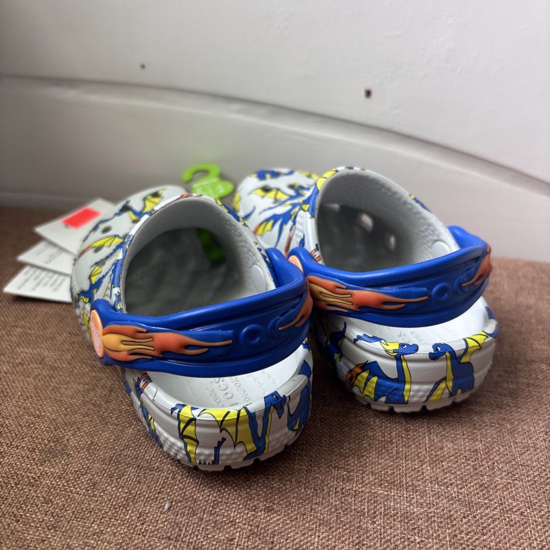 Crocs Classic Dragon Graphic Clog Toddler Size C6 (Category: Babies & Kids Fashion) located in Quezon City. Submitted by eshymarie (Listing ID 1470168491, Image 3). Description: - Item: Crocs Classic Dragon Graphic Clog for Toddlers - Size: C6 (EUR 22-23) for 13cm foot length - Design: White base with vibrant blue and yellow dragon print - Features: Blue pivoting heel strap with flame details on the sides - Condition: Brand new with original tags