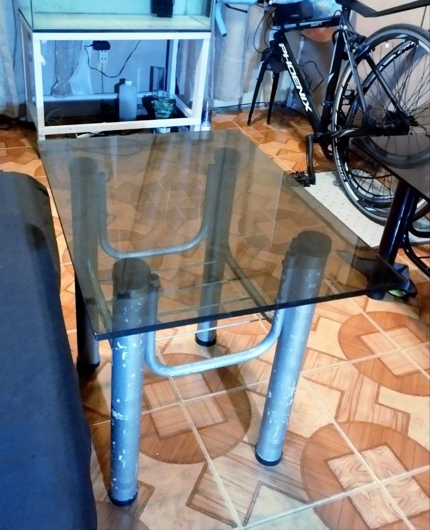 Glass Top Metal Frame Coffee Table With Lower Shelf (Category: Tables & Sets) located in Mandaluyong City. Submitted by perfectfashion.mnl (Listing ID 1470288966, Image 3). Description: - Glass top coffee table with a sturdy metal tubular frame - Dimensions: 24 inches x 21 inches - Features a lower wire shelf for extra storage - Perfect for living rooms, bedrooms, or small apartments - Condition: Silver painted legs show paint chipping and wear a