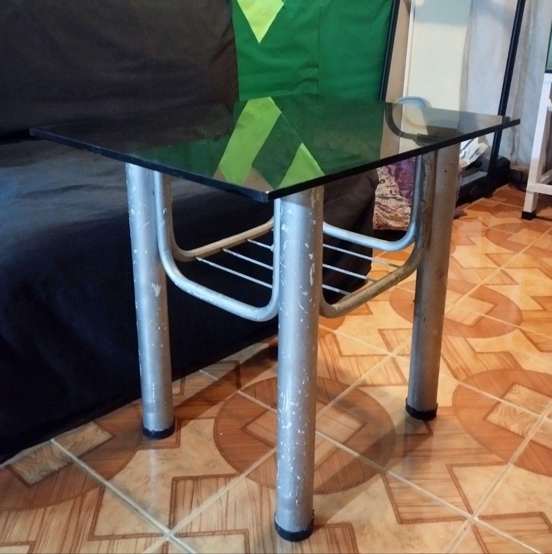 Glass Top Metal Frame Coffee Table With Lower Shelf (Category: Tables & Sets) located in Mandaluyong City. Submitted by perfectfashion.mnl (Listing ID 1470288966, Image 2). Description: - Glass top coffee table with a sturdy metal tubular frame - Dimensions: 24 inches x 21 inches - Features a lower wire shelf for extra storage - Perfect for living rooms, bedrooms, or small apartments - Condition: Silver painted legs show paint chipping and wear a