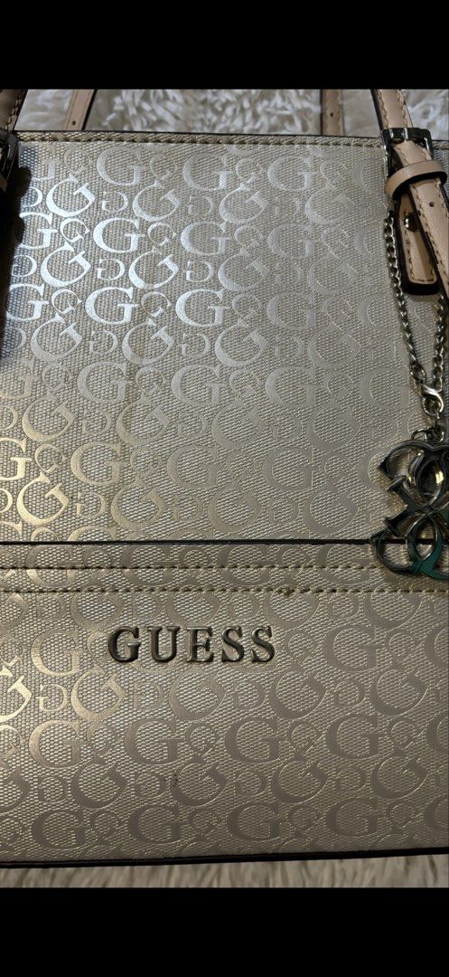 Guess Metallic Gold Monogram Tote Bag (Category: Luxury Bags & Wallets) located in Rizal. Submitted by kianapreloves (Listing ID 1470212396, Image 2). Description: - Brand: Guess - Color: Metallic gold with tan leather-look handles and trim - Pattern: All-over monogram print - Hardware: Metal Guess logo on the front and a stylish hanging logo charm - Closure: Secure zip top closure - Interior: Fabric monogram-patterned lining with an interior pla