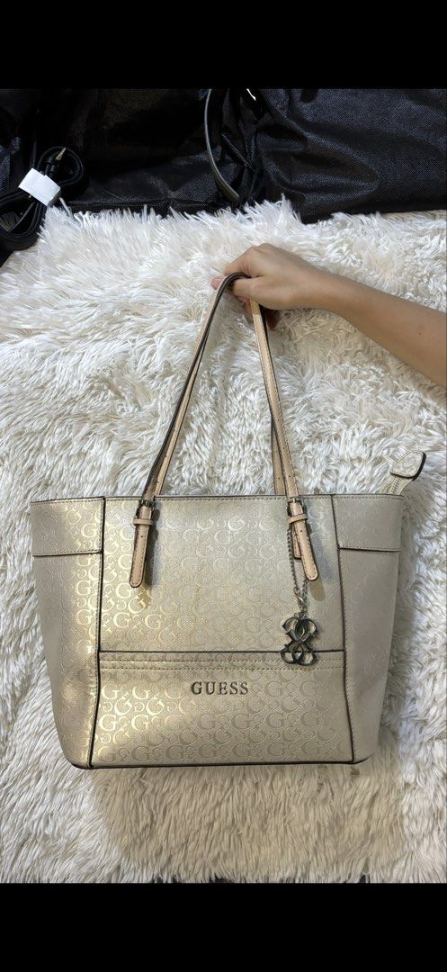 Guess Metallic Gold Monogram Tote Bag (Category: Luxury Bags & Wallets) located in Rizal. Submitted by kianapreloves (Listing ID 1470212396, Image 1). Description: - Brand: Guess - Color: Metallic gold with tan leather-look handles and trim - Pattern: All-over monogram print - Hardware: Metal Guess logo on the front and a stylish hanging logo charm - Closure: Secure zip top closure - Interior: Fabric monogram-patterned lining with an interior pla