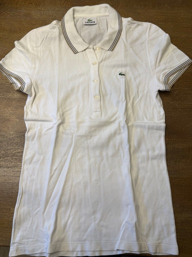 Lacoste White Polo Shirt Size 38 (Category: Shirts) located in Muntinlupa City. Submitted by el1b12345 (Listing ID 1470186067, Image 1). Description: - White Lacoste polo shirt in women's - size 38. - L - 22" - W - 17" - Striped collar and sleeve cuffs in brown, black, and gold tones. - Classic fit with four-button placket and embroidered crocodile logo on the chest. - Cotton petit pique fabric. - Clean condition, no major flaws found. - mop: Gca