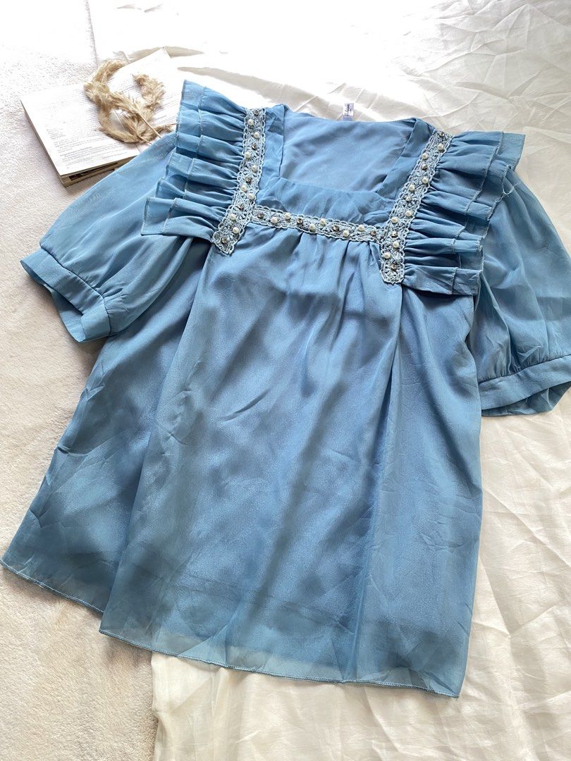 Light blue ruffle blouse with pearl trim Medium 37 (Category: Blouses) located in Poro. Submitted by joyceyuu (Listing ID 1470169049, Image 2). Description: - Color: Light blue with a subtle sheen - Style: Loose fit, perfect for casual or dressed-up looks - Neckline: Square neck with elegant pearl and lace detailing - Details: Eye-catching ruffles across the front shoulders - Sleeves: Short puff sleeves with comfortable gathered cuffs - Material: