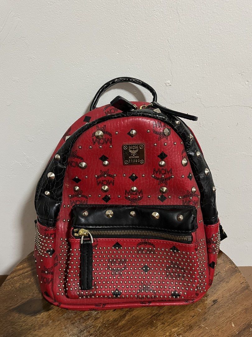 MCM Red Studded Leather Backpack (Category: Backpacks) located in Cabuyao City. Submitted by rdtrmt (Listing ID 1470151903, Image 1). Description: - Brand: MCM - Color: Bold red with black leather trim and silver-tone hardware - Design: Classic MCM monogram print covered in stylish metal studs - Features: Front zippered pocket with hardware pull, top grab handle, and signature front logo plaque - Straps: Adjustable black fabric shoulder straps fo