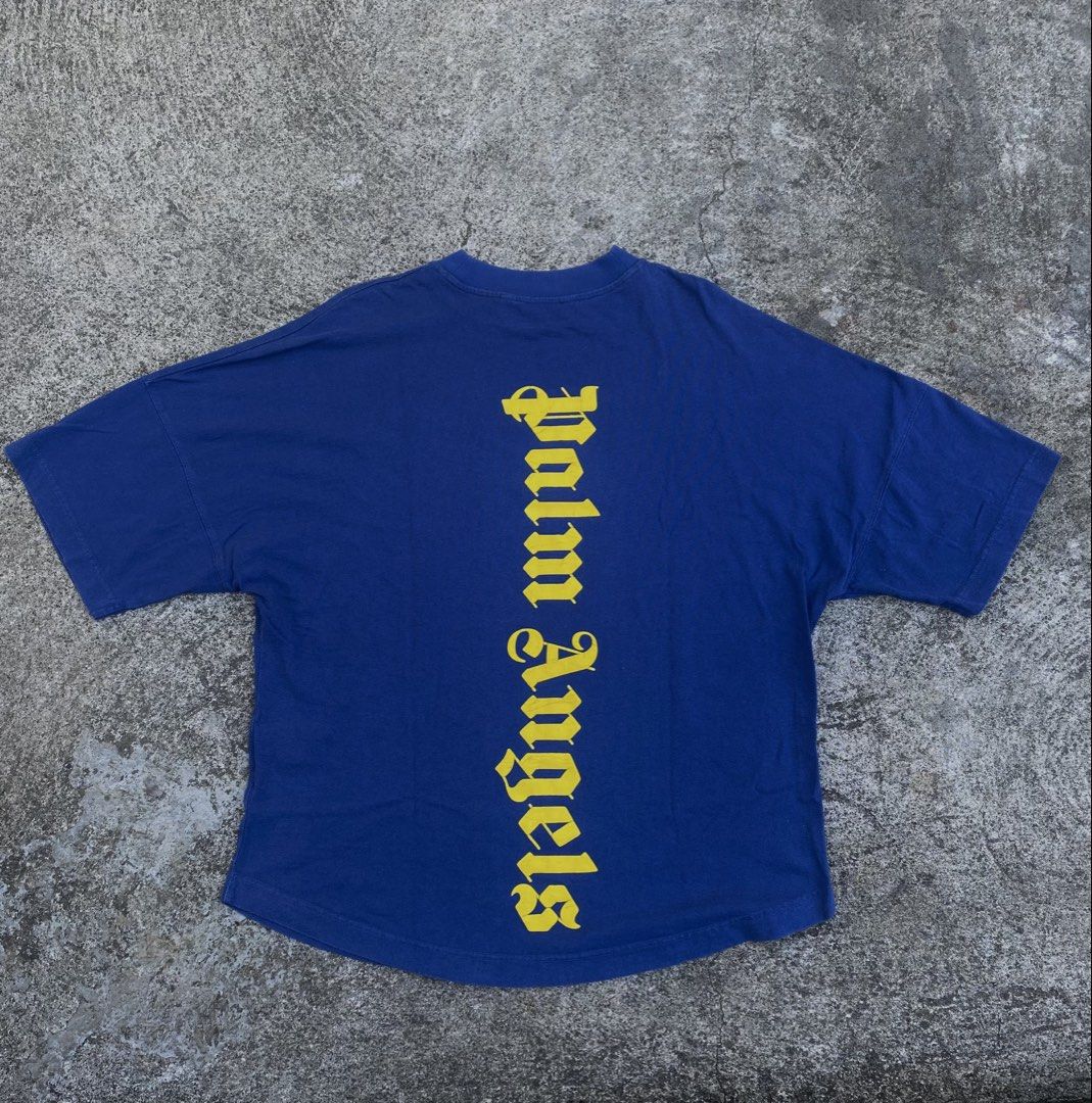 Palm Angels Blue Yellow Logo Tee (Category: Tshirts & Polo Shirts ) located in Iba. Submitted by gotmethrift (Listing ID 1470295193, Image 3). Description: - Brand: Palm Angels - Color: Royal blue with yellow gothic logos - Design: Small front neckline logo and giant vertical back logo - Fit: Oversized with drop shoulders and curved hem - Material: Heavyweight cotton - Style: Streetwear, pairs easily with jeans or cargos.