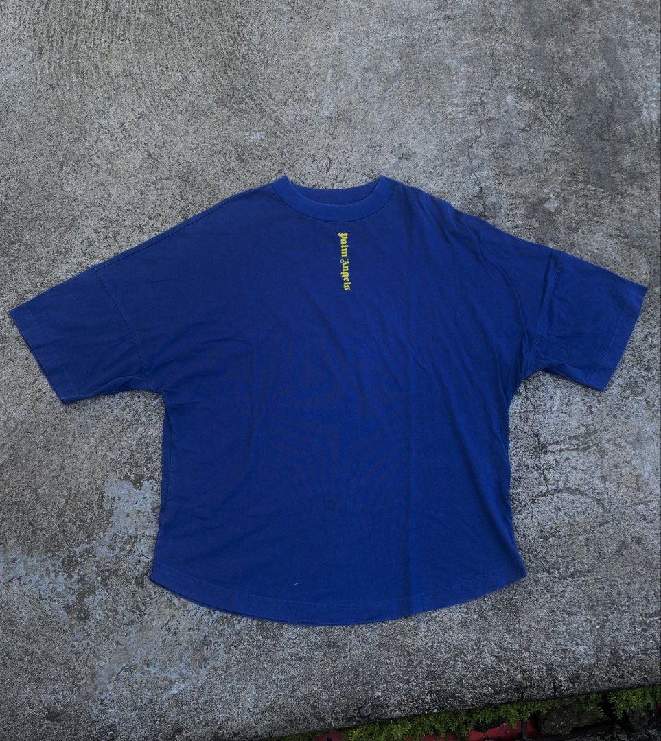 Palm Angels Blue Yellow Logo Tee (Category: Tshirts & Polo Shirts ) located in Iba. Submitted by gotmethrift (Listing ID 1470295193, Image 1). Description: - Brand: Palm Angels - Color: Royal blue with yellow gothic logos - Design: Small front neckline logo and giant vertical back logo - Fit: Oversized with drop shoulders and curved hem - Material: Heavyweight cotton - Style: Streetwear, pairs easily with jeans or cargos.