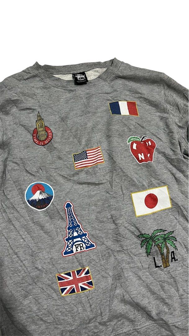 Stüssy Grey Patchwork Crewneck Sweatshirt (Category: Coats, Jackets and Outerwear) located in Valencia. Submitted by thriftyfinds01 (Listing ID 1470252779, Image 3). Description: Stüssy grey crewneck sweatshirt covered in embroidered city and country patches. Front has flags, Eiffel Tower, Big Ben, and Fuji. Back features Brooklyn Bridge, California wave, surf board, and parental advisory patch. Ribbed cuffs and hem. Stüssy neck tag present. Size