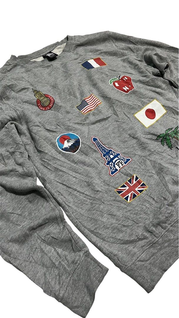 Stüssy Grey Patchwork Crewneck Sweatshirt (Category: Coats, Jackets and Outerwear) located in Valencia. Submitted by thriftyfinds01 (Listing ID 1470252779, Image 2). Description: Stüssy grey crewneck sweatshirt covered in embroidered city and country patches. Front has flags, Eiffel Tower, Big Ben, and Fuji. Back features Brooklyn Bridge, California wave, surf board, and parental advisory patch. Ribbed cuffs and hem. Stüssy neck tag present. Size
