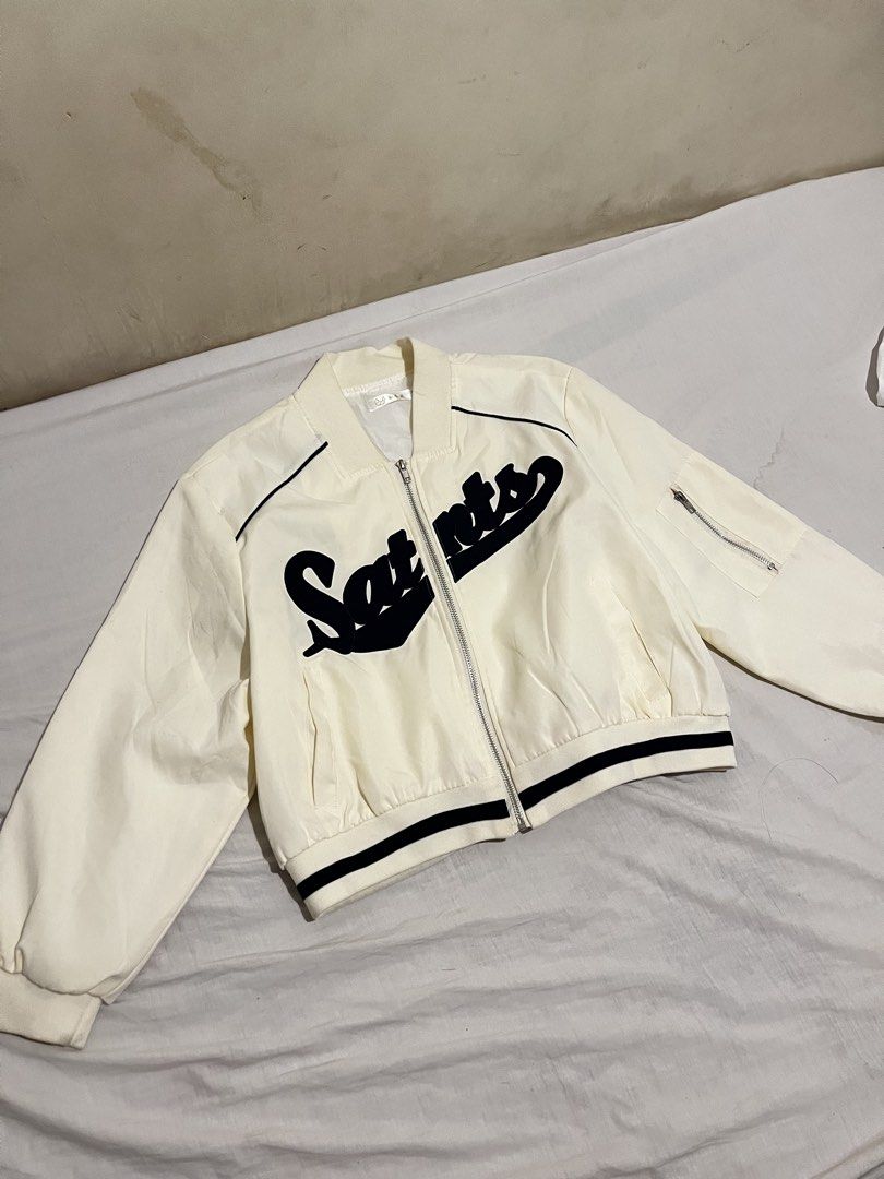 White and Black Saints Letterman Bomber Jacket (Category: Coats, Jackets and Outerwear) located in San Fernando. Submitted by angelhave_wings (Listing ID 1470291101, Image 3). Description: - Color: White with bold black 'Saints' lettering on the chest - Closure: Zip-up front - Pockets: Convenient side pockets and a utility pocket on the left sleeve - Details: Ribbed cuffs and hem with stylish black contrast striping - Fit: Lightweight style, idea