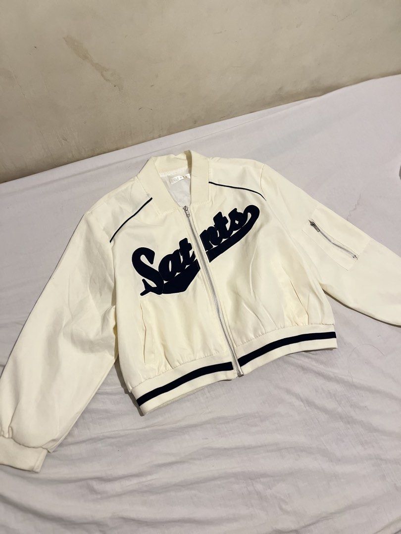 White and Black Saints Letterman Bomber Jacket (Category: Coats, Jackets and Outerwear) located in San Fernando. Submitted by angelhave_wings (Listing ID 1470291101, Image 1). Description: - Color: White with bold black 'Saints' lettering on the chest - Closure: Zip-up front - Pockets: Convenient side pockets and a utility pocket on the left sleeve - Details: Ribbed cuffs and hem with stylish black contrast striping - Fit: Lightweight style, idea