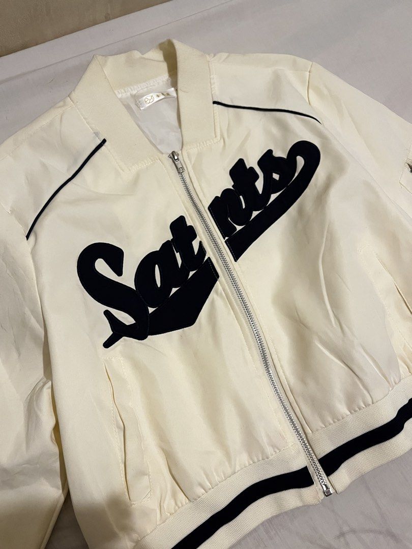 White and Black Saints Letterman Bomber Jacket (Category: Coats, Jackets and Outerwear) located in San Fernando. Submitted by angelhave_wings (Listing ID 1470291101, Image 2). Description: - Color: White with bold black 'Saints' lettering on the chest - Closure: Zip-up front - Pockets: Convenient side pockets and a utility pocket on the left sleeve - Details: Ribbed cuffs and hem with stylish black contrast striping - Fit: Lightweight style, idea
