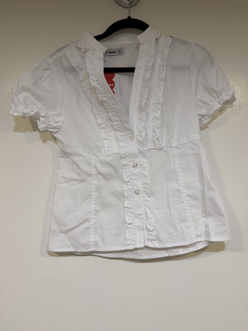 White ruffle button-up short sleeve blouse top (Category: Blouses) located in Mandaluyong City. Submitted by closet.me (Listing ID 1470277755, Image 1). Description: - Brand new with original tag attached - Color: Solid white - Material: Lightweight cotton-blend - Design: Ruffled front placket, high collar with button closure - Sleeves: Short puff sleeves with ruffled cuffs - Condition: Pristine, never worn.