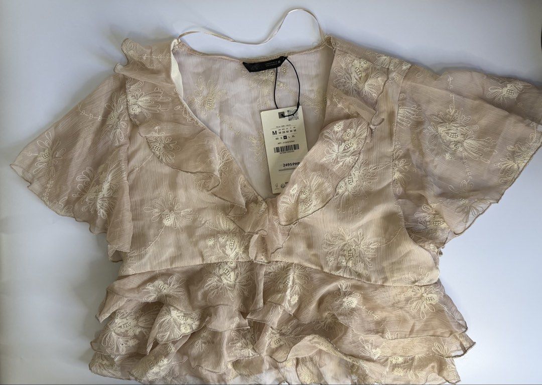 Zara Embroidered Ruffle Blouse Beige Size M (Category: Blouses) located in General Santos City (Dadiangas). Submitted by my_lzlove (Listing ID 1470241470, Image 1). Description: - Brand: Zara - Size: Medium (Brand new with original tags) - Original Price: ₱2,495 - Color: Beige with delicate white floral embroidery - Details: Deep V-neckline with ruffled trim, short flutter sleeves, and layered tiered ruffle hem - Fabric: Lightweight and textured 